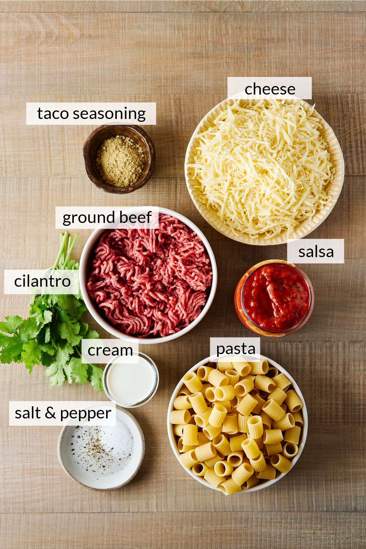 Bowl of ground beef near bowls of cheese, salsa, taco seasoning and uncooked pasta.