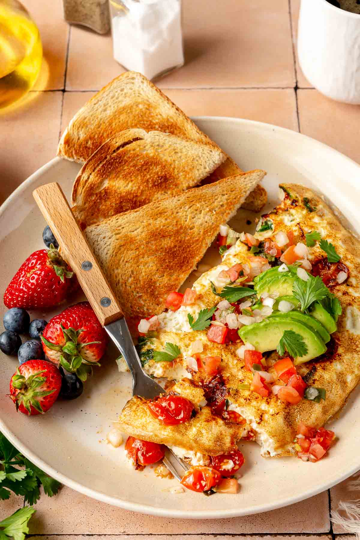 A fork cutting into an omelette on a plate with toast and fruit.