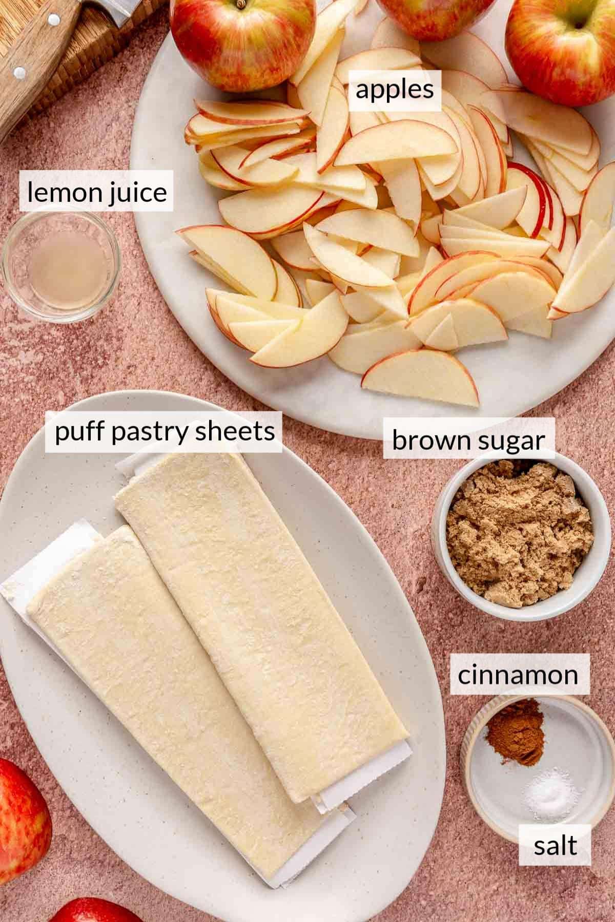 Sliced apples on a white plate near sheets of puff pastry and a bowl of brown sugar.