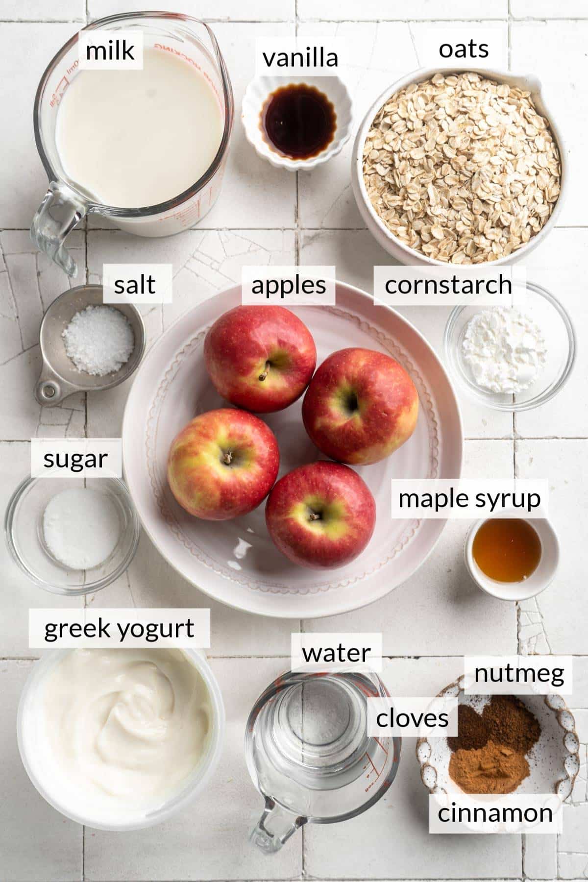 Bowl filled with apples near bowls of milk, oats, vanilla, greek yogurt, spices, water and cornstarch.