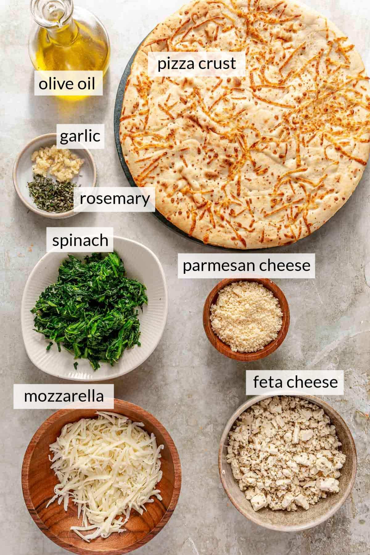 Pizza crust near small bowls of cheese, spinach, garlic and rosemary.