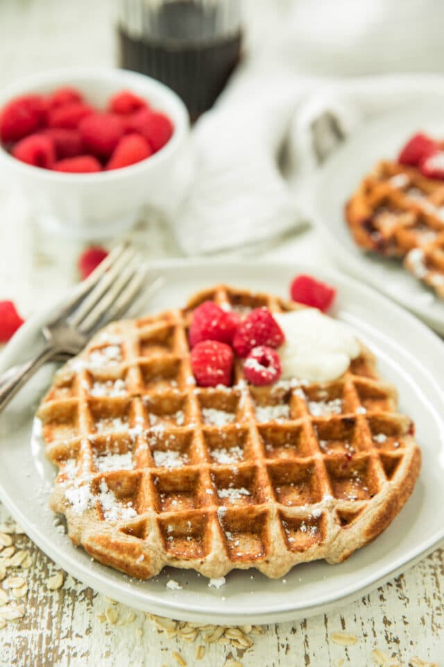 side view of Healthy Raspberry Banana Oatmeal Greek Yogurt Waffles on white plates with a small white bowl of red raspberries