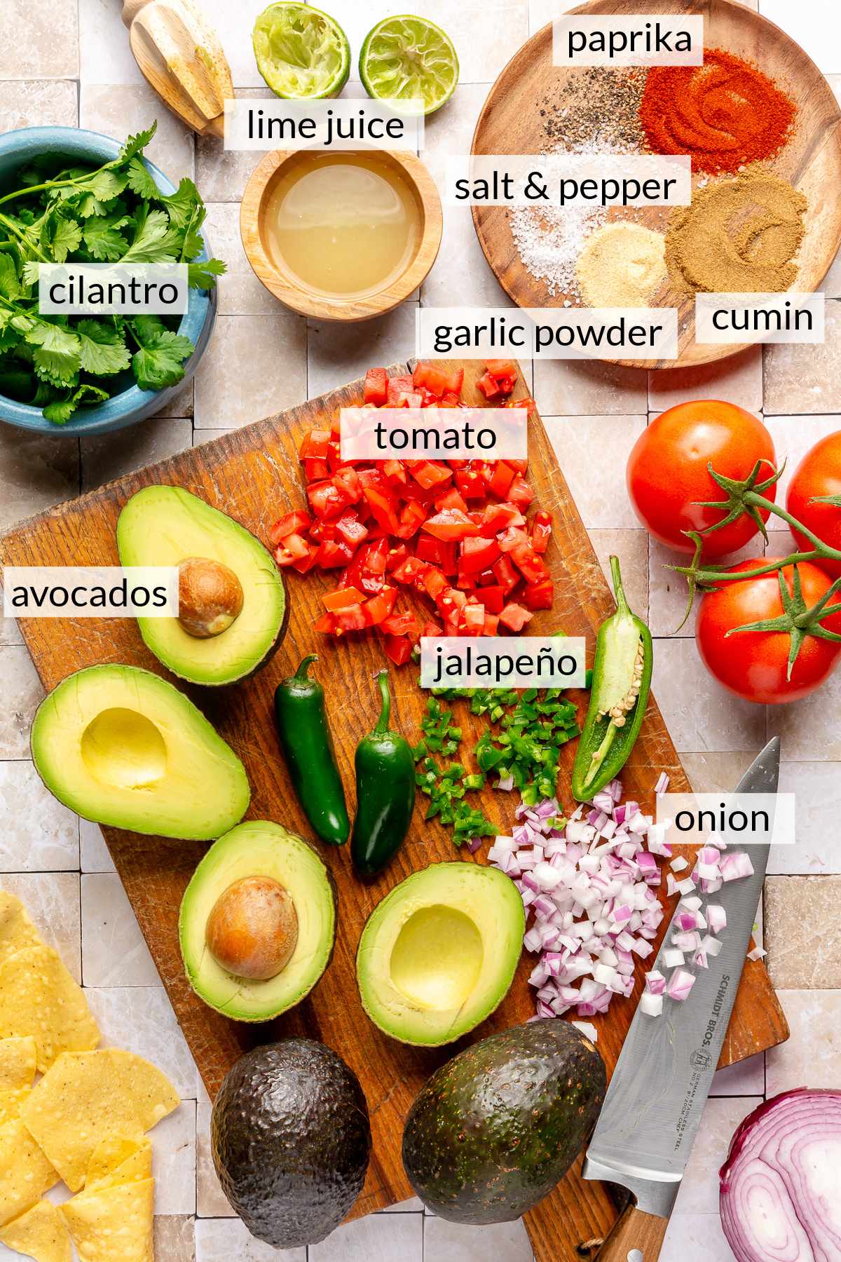 Avocados cut in half, diced tomato, diced onion and jalapeños on a cutting board.