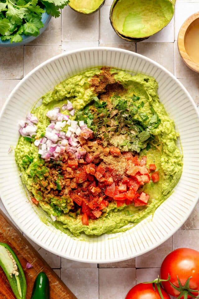 Spices, diced tomato and diced onion in a bowl with mashed avocado.