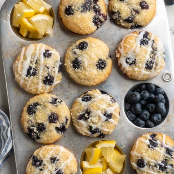 Lemon blueberry muffins in a muffin pan with fresh blueberries and lemon wedges.