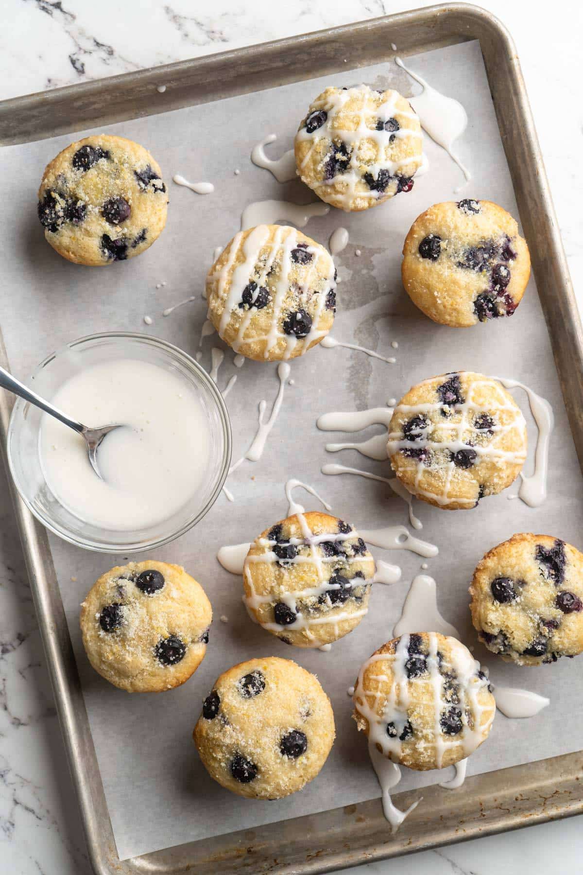 Drizzling glaze over lemon blueberry muffins on a cookie sheet.