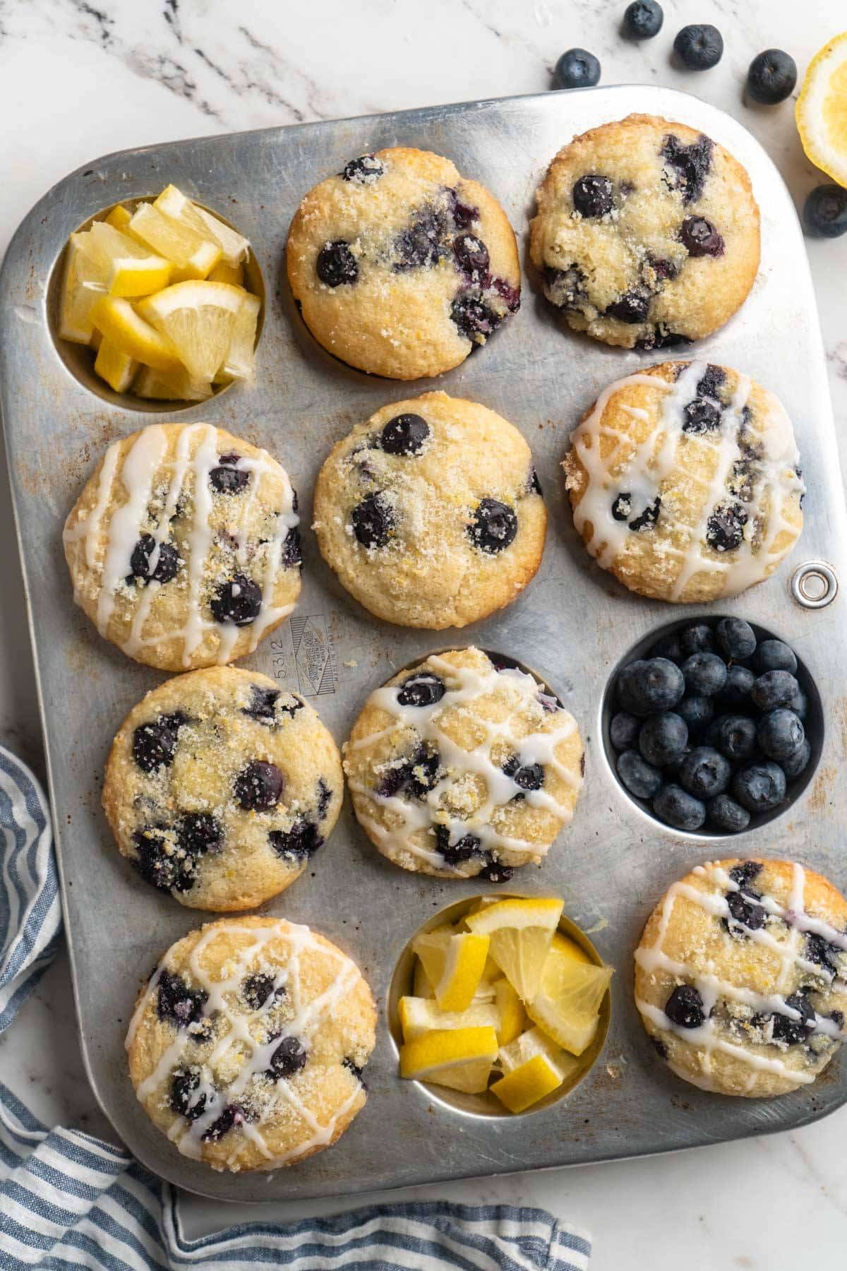 Lemon blueberry muffins in a muffin pan with fresh blueberries and lemon wedges.