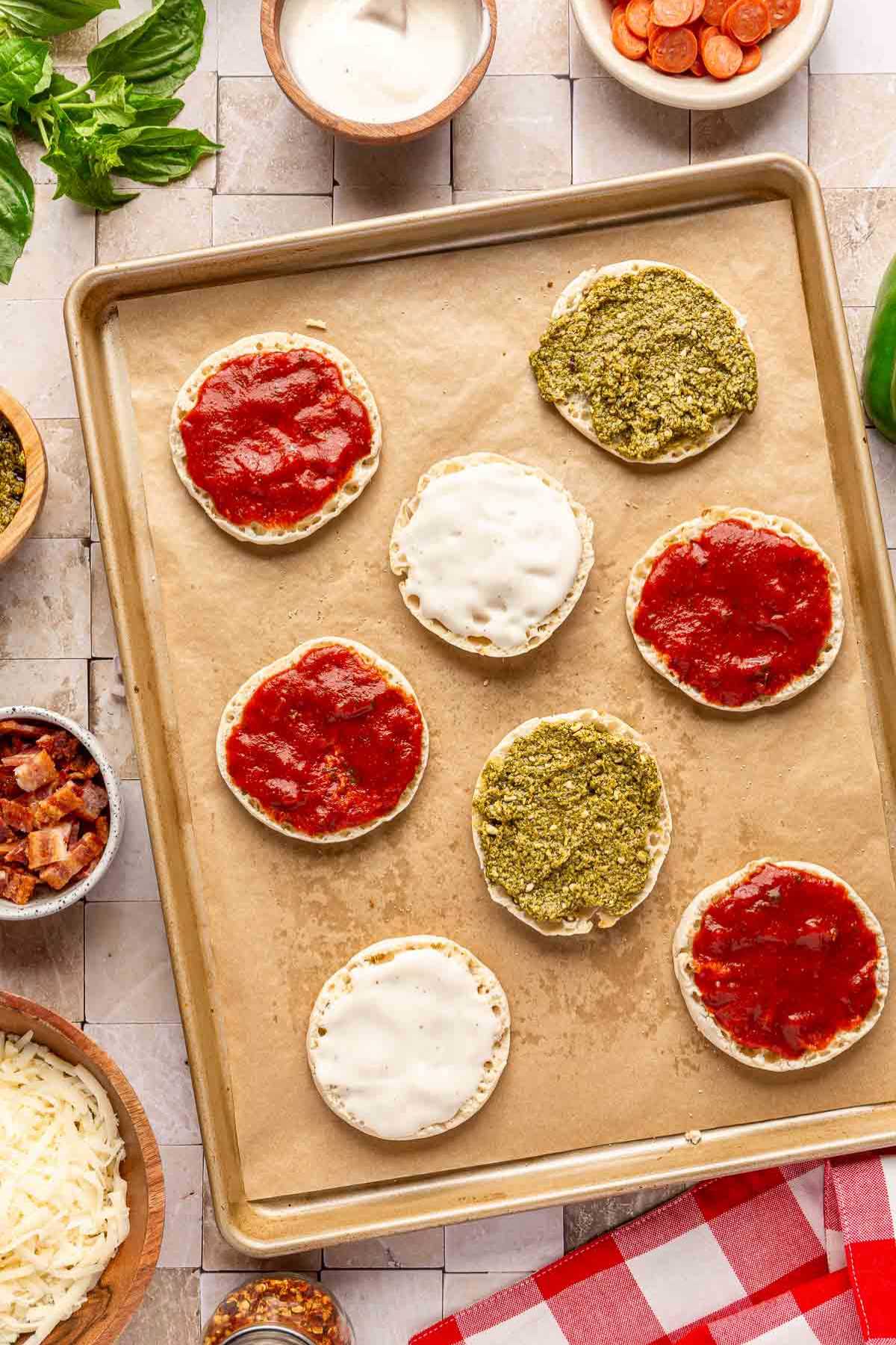 Sauce spread on the top of English muffin halves on a baking sheet.