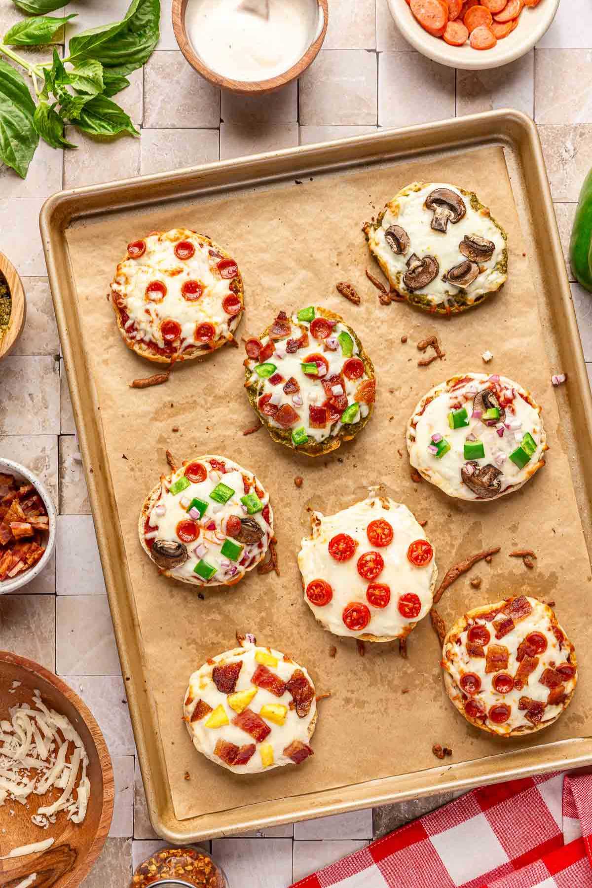 Baked English muffin pizzas with different toppings on a pan.