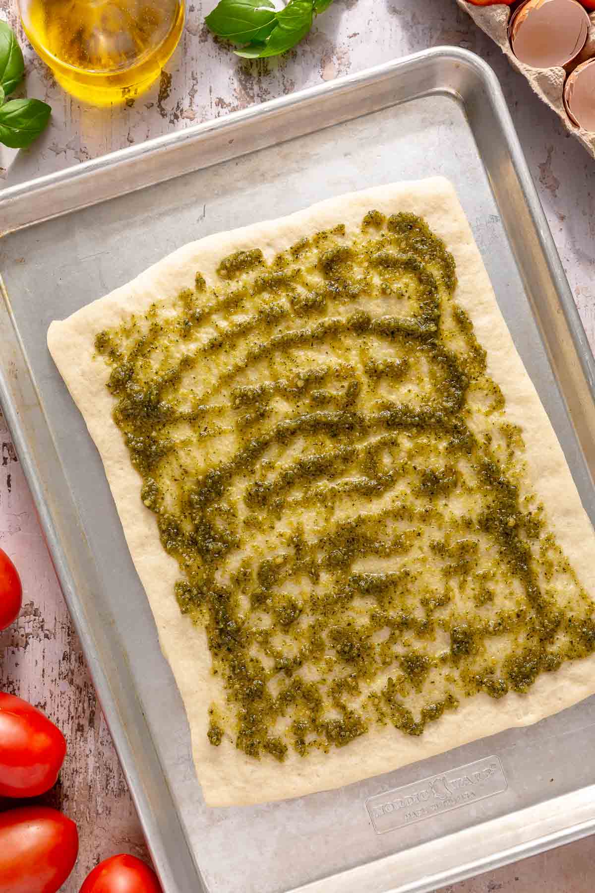 Pesto spread over pizza dough on a baking sheet pan.