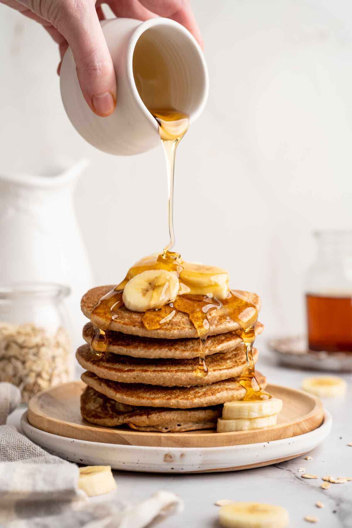 Hand pouring syrup over a stack of banana oatmeal pancakes.