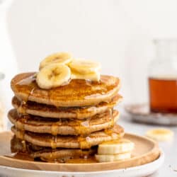 A stack of banana oatmeal pancakes topped with banana slices, butter and maple syrup.