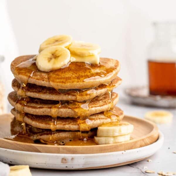 A stack of banana oatmeal pancakes topped with banana slices, butter and maple syrup.