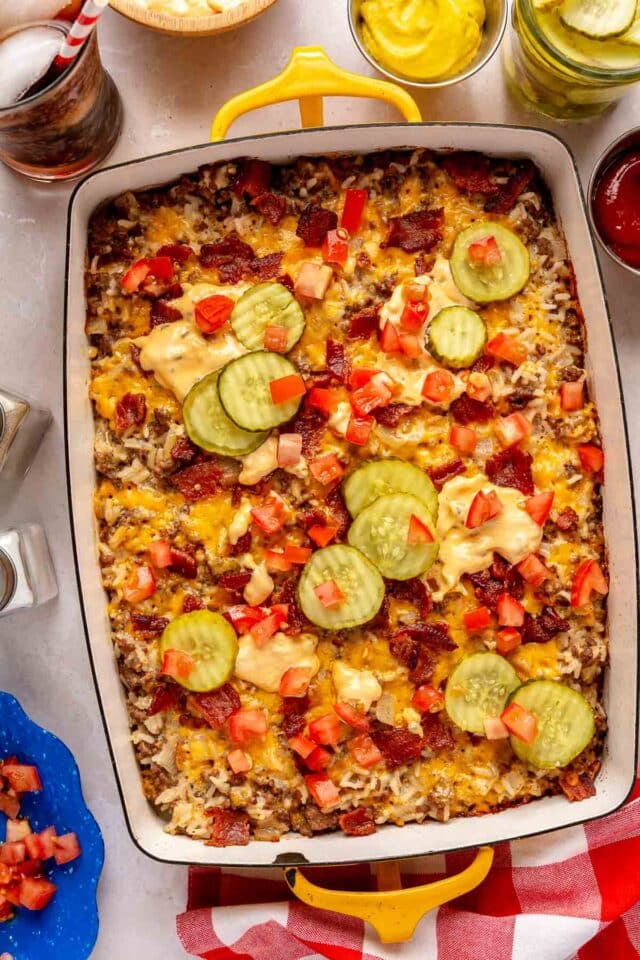 Cheeseburger casserole in a dish served near a glass of soda, mustard and pickles.