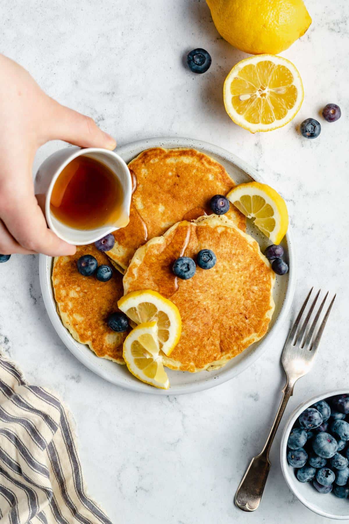 Drizzling pancake syrup over a plate full of pancakes topped with blueberries.