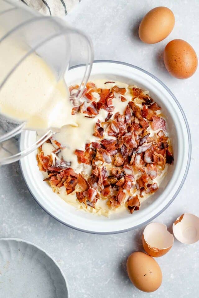 Pouring milk mixture over bacon and hash browns in a pie plate.