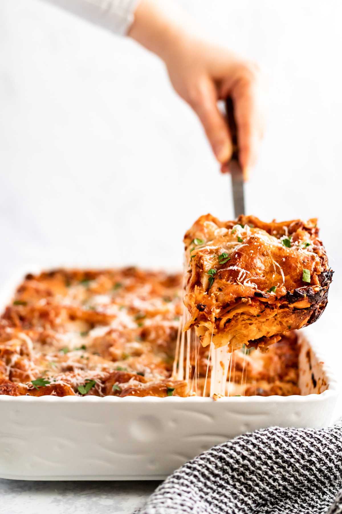 Woman's hand scooping a serving of lasagna out of a large white dish.