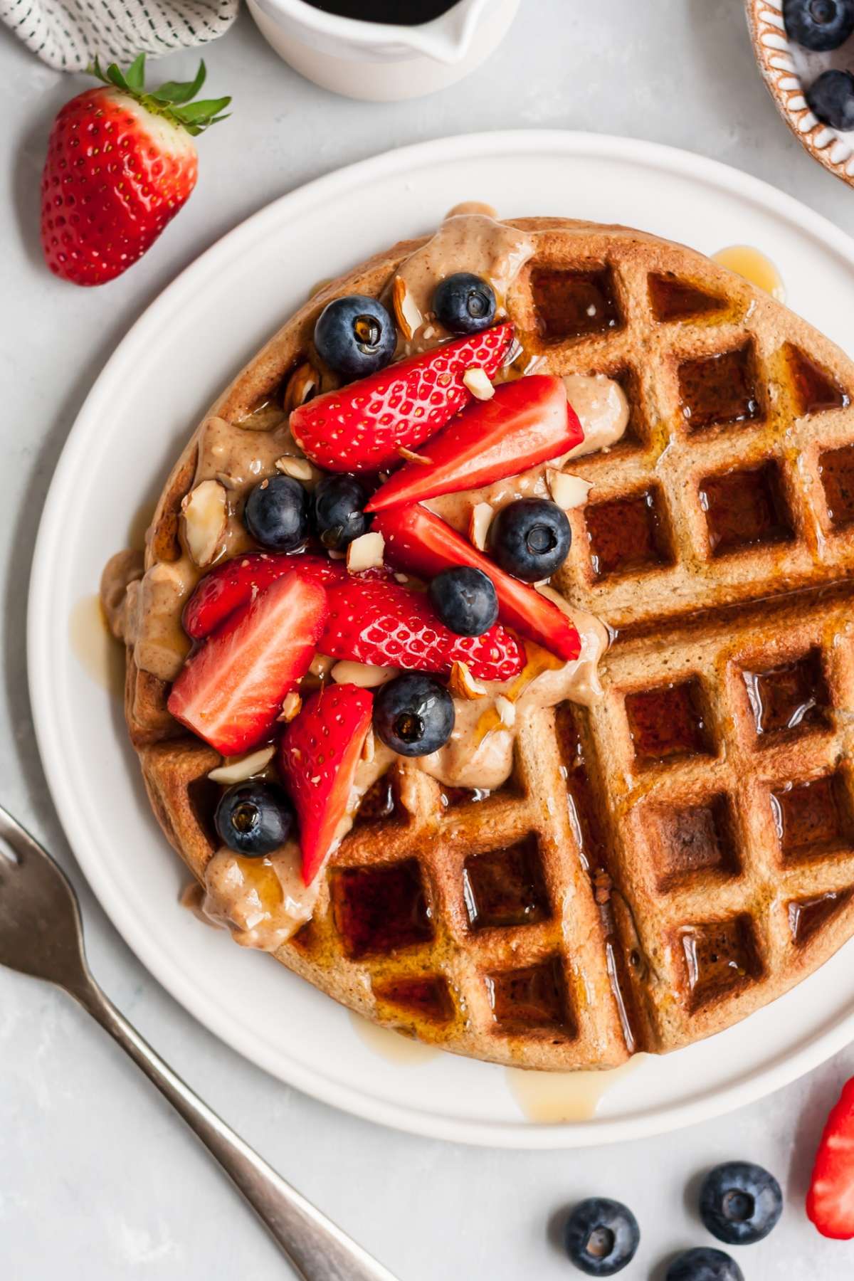 Large waffle on a white plate with strawberries, blueberries, maple syrup and almond butter on top.