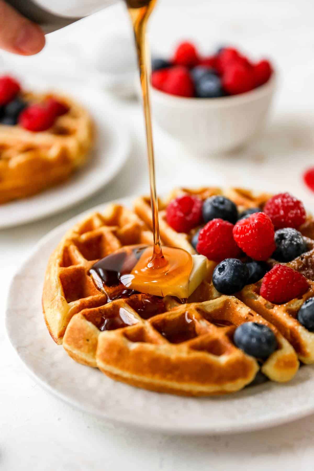 Drizzling maple syrup over a fluffy waffle on a white plate.