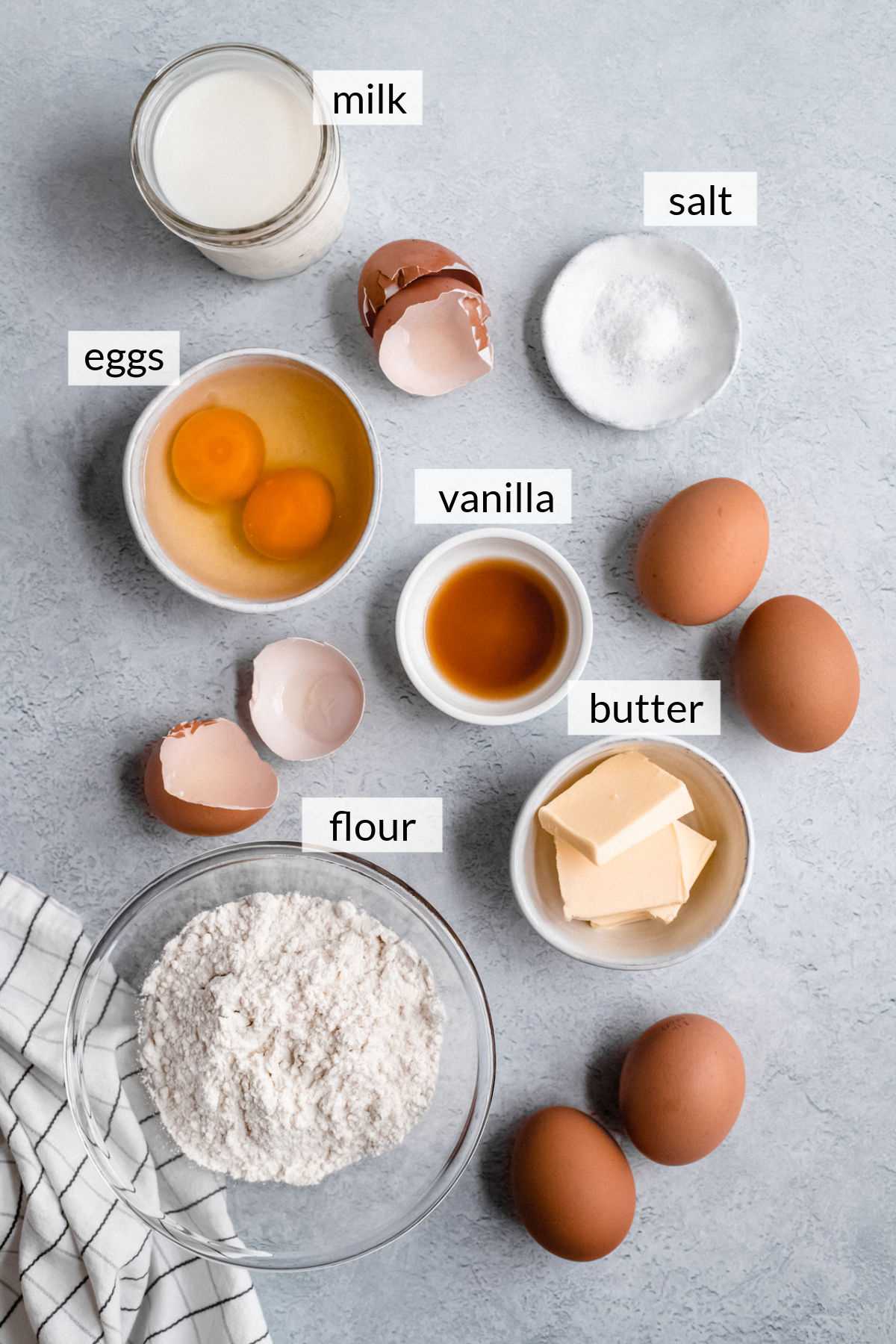 Bowl of flour, butter, eggs, milk, salt and vanilla near cracked egg shells.