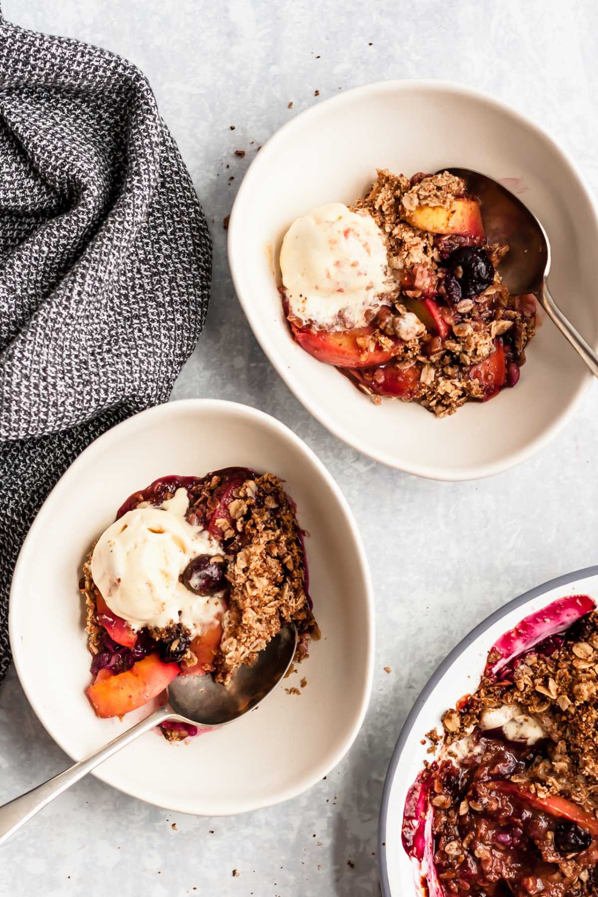 Servings of peach blueberry crisp divided into white bowls and served with a scoop of vanilla ice cream.