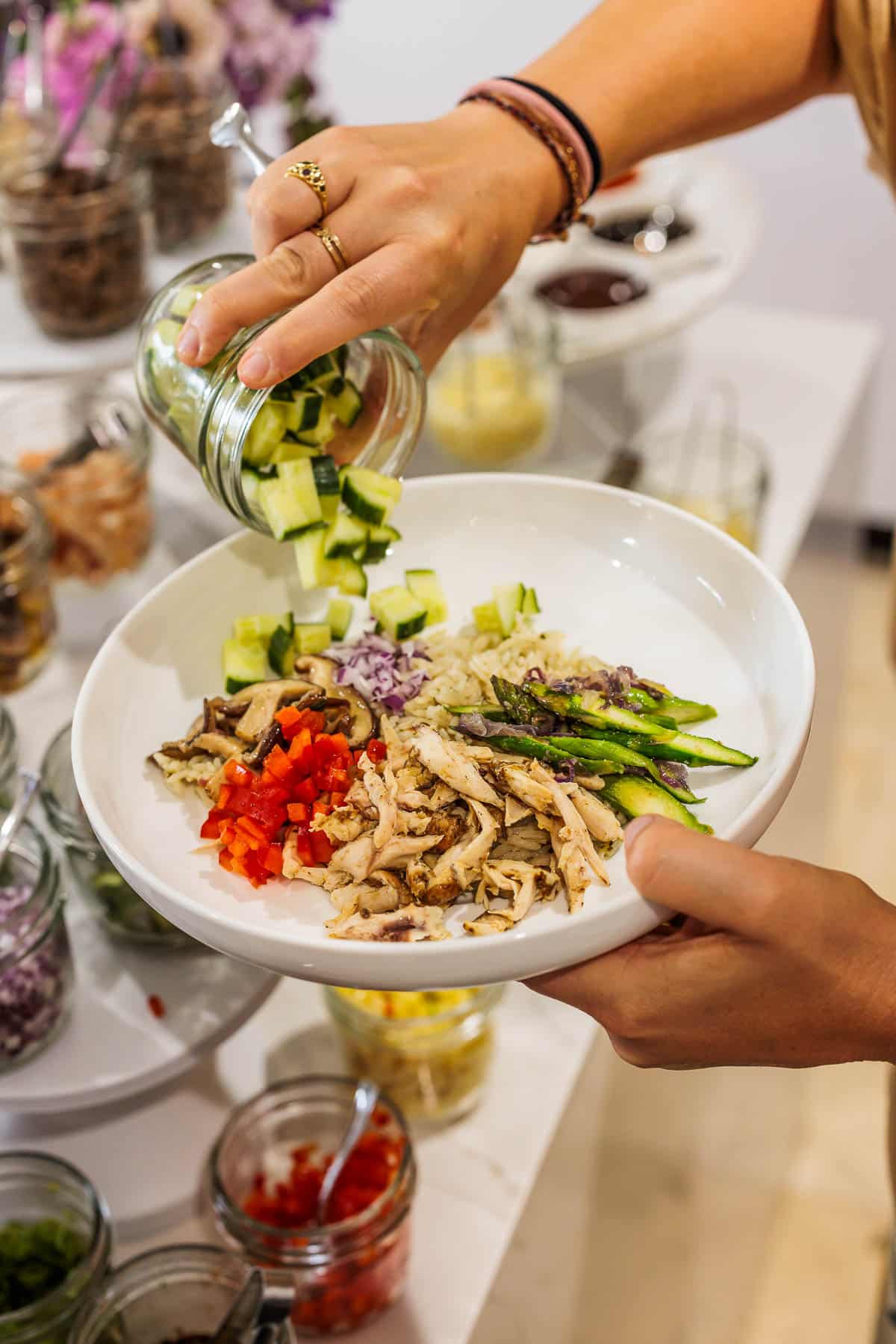 Pouring out diced cucumbers from a small mason jar into a bowl with rice and vegetables.