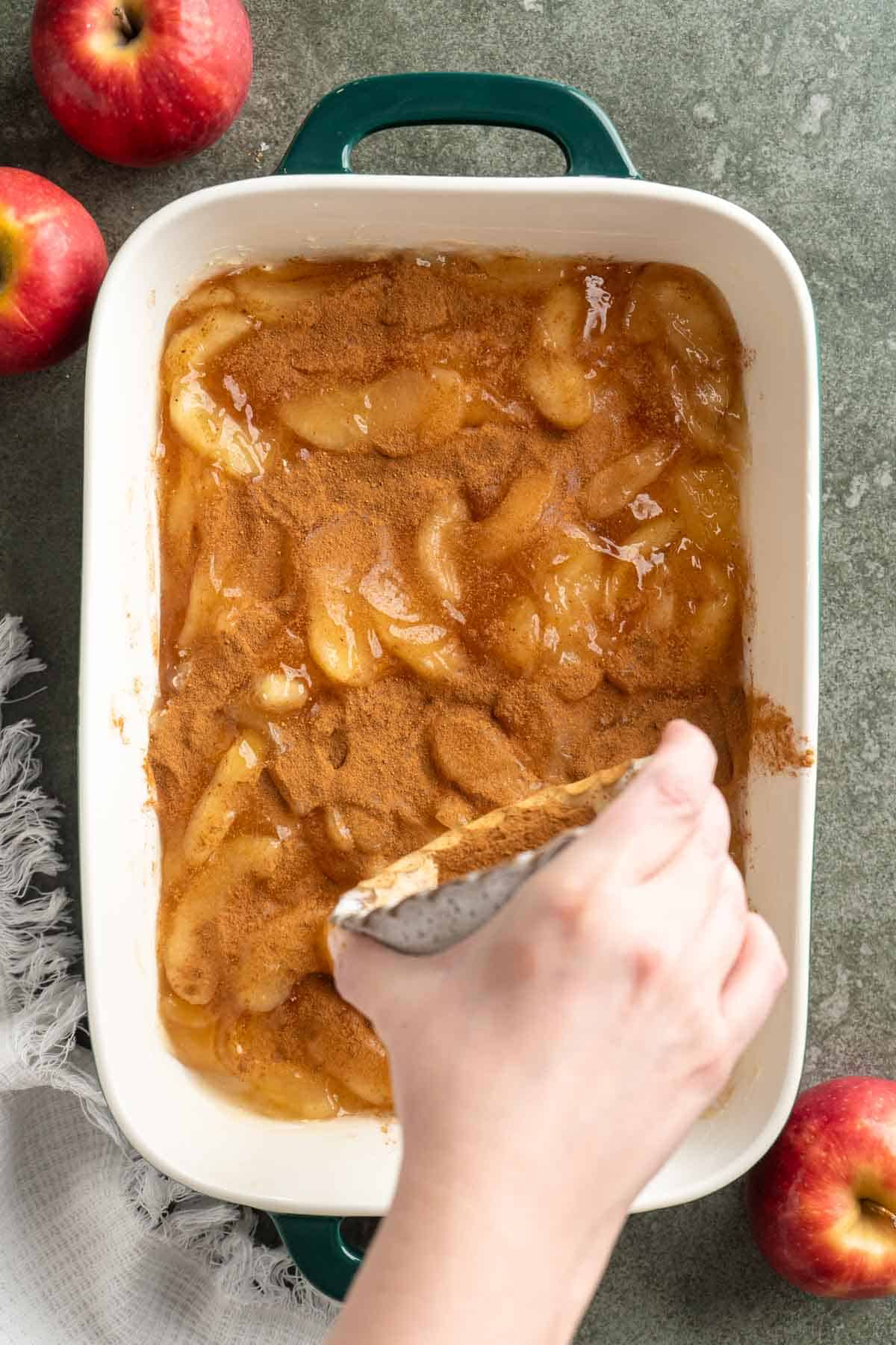 Hand holding a small bowl of spices over sliced apples and sprinkling the spices over the top.