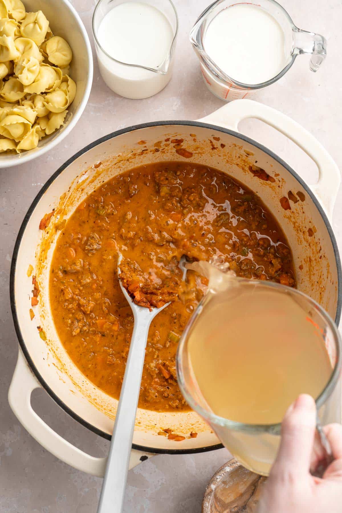 Pouring chicken broth into a pot with Italian sausage, veggies and tomato paste.