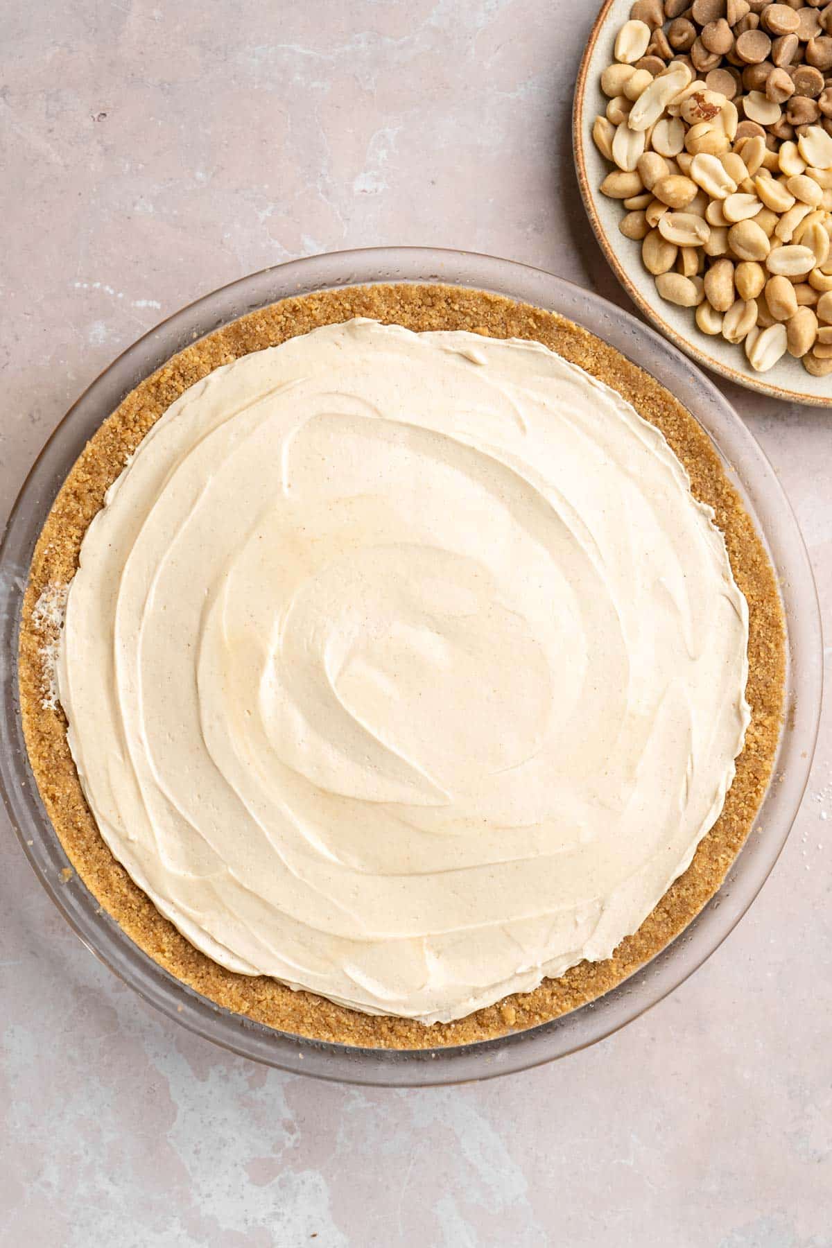 Peanut butter filling in a graham cracker crust near a bowl of chopped peanuts.